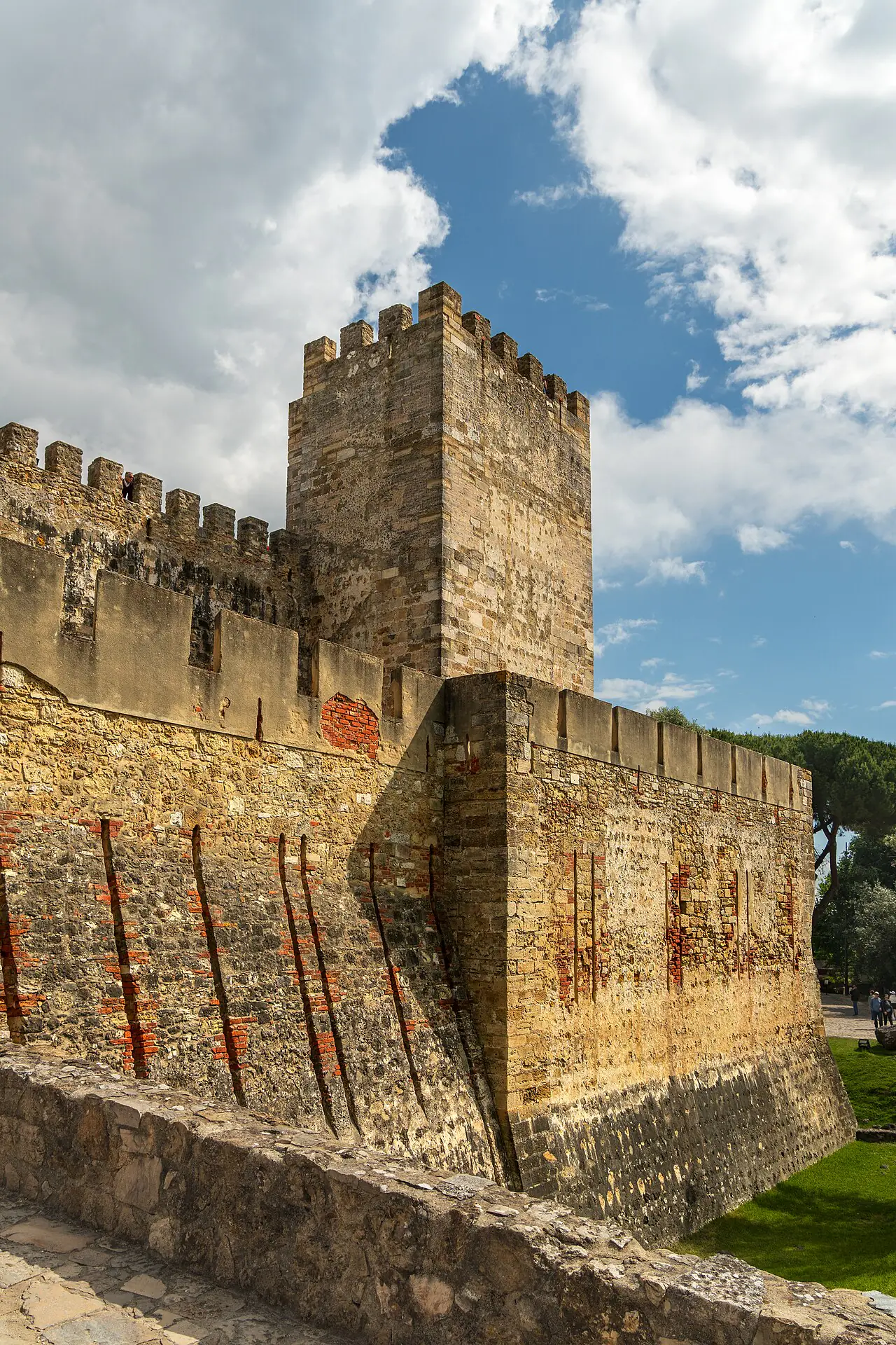 A stone tower and crenellated walls at São Jorge Castle in warm sunlight beneath a cloud-filled sky.