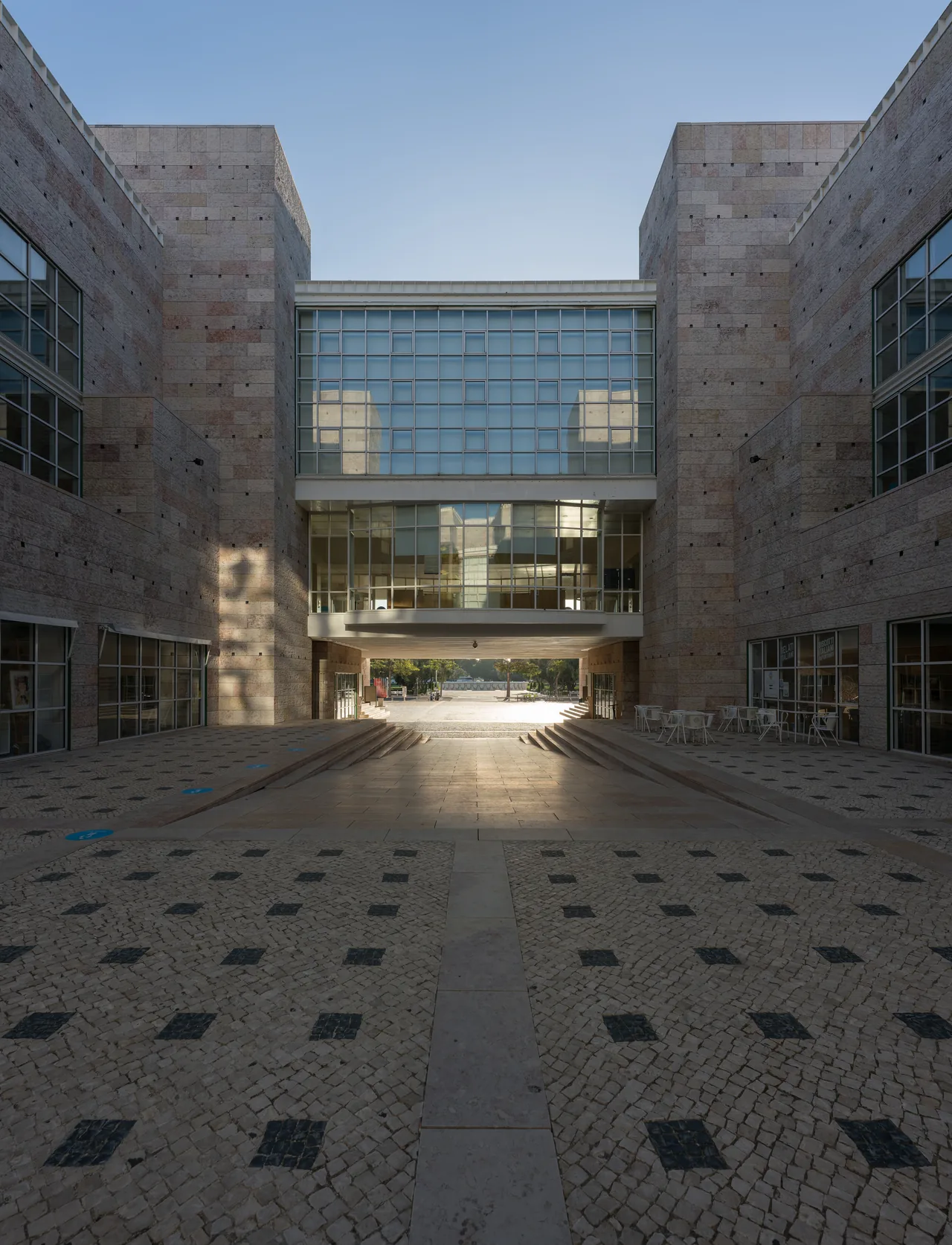 Paredes altas de pedra a enquadrar um pátio pavimentado e uma passagem de vidro no Centro Cultural de Belém.