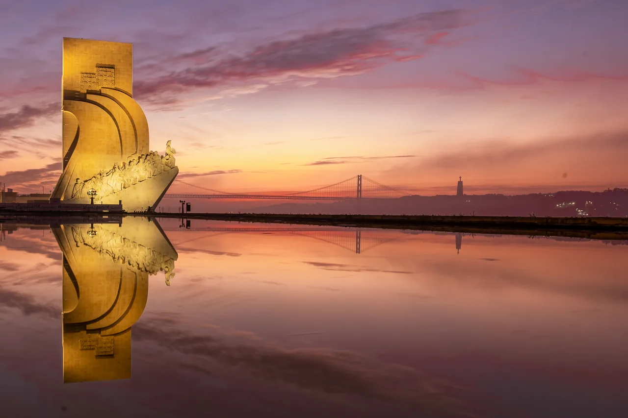 The Monument to the Discoveries reflected in shallow water beneath a pink and orange sunrise.