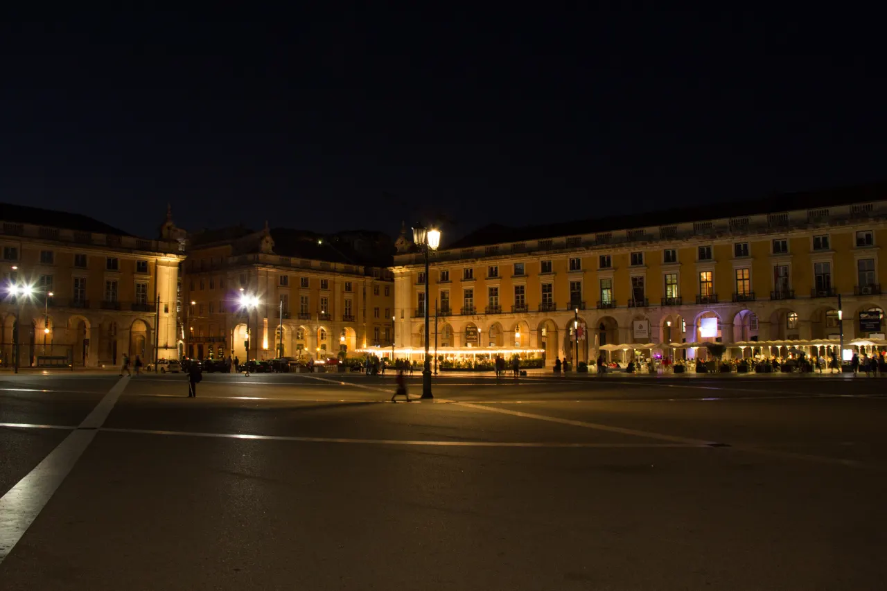 Illuminated yellow arcades and café terraces around Praça do Comércio at night.